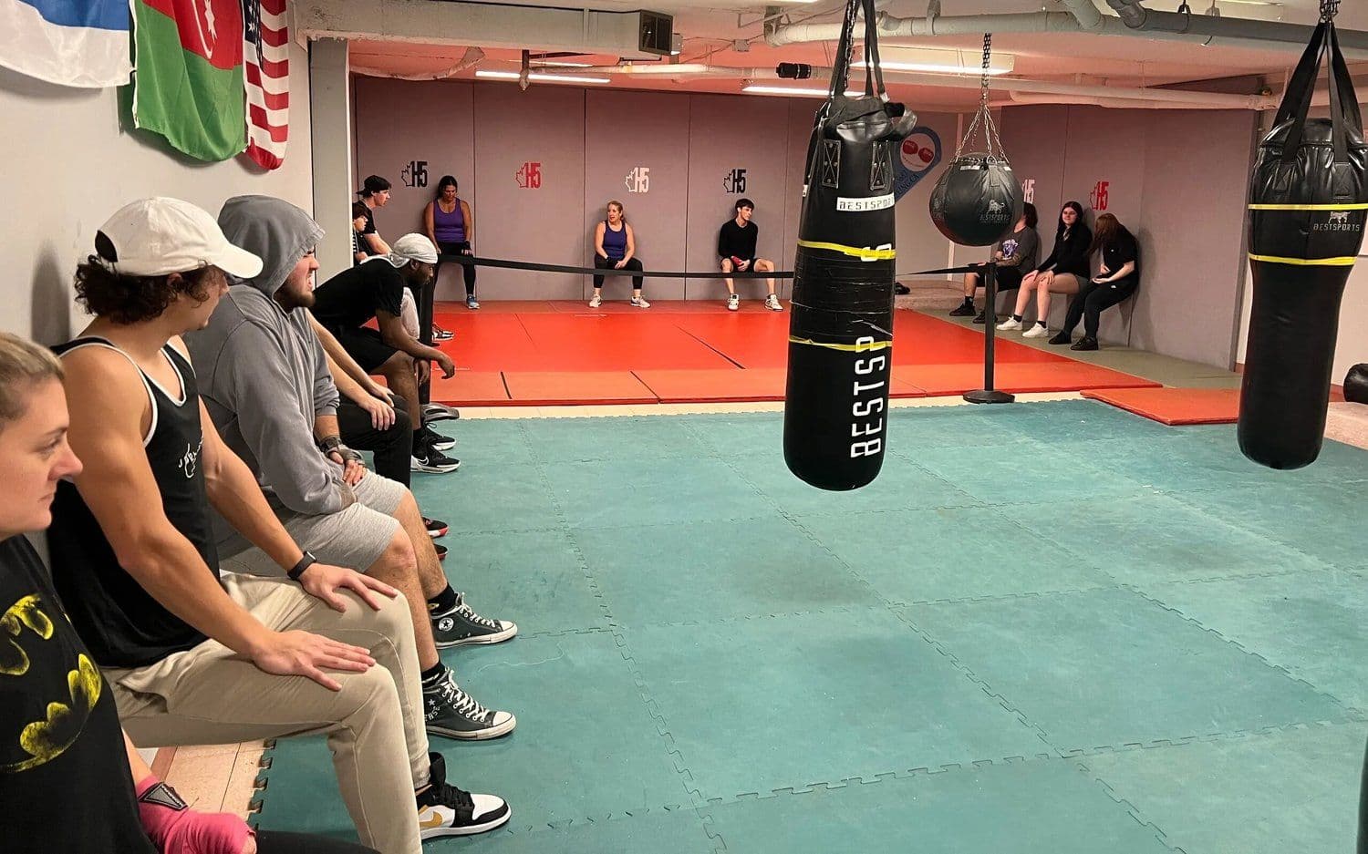 A boxing class seated along the wall of a training room with heavy bags